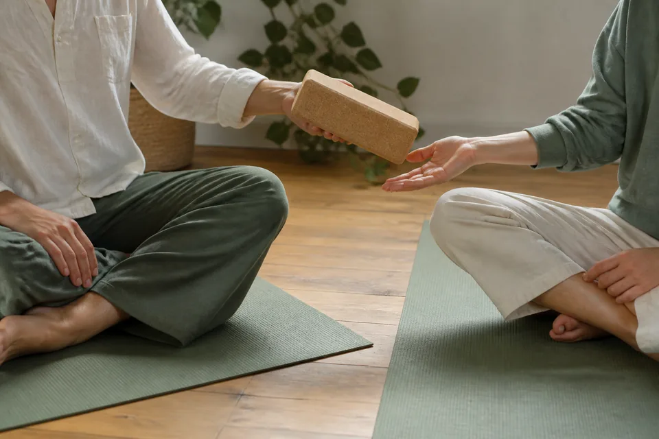 A cork yoga block passed between two people seated on mats.